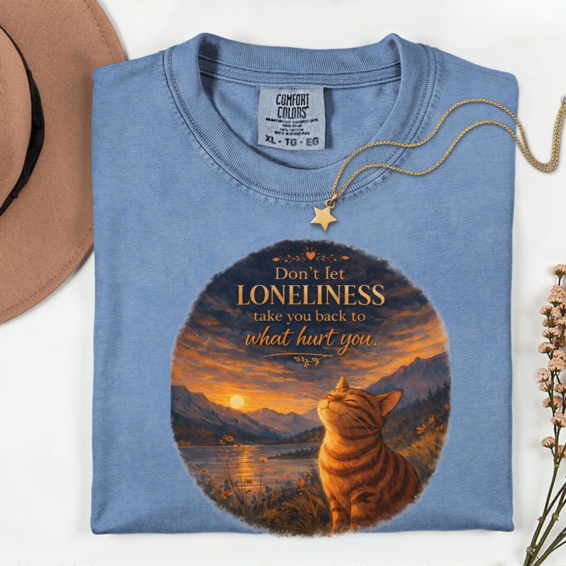 May include: A light blue Comfort Colors t-shirt with a graphic of a cat and a sunset scene. The text reads "Don't let LONELINESS take you back to what hurt you." A gold star pendant necklace is draped across the shirt.