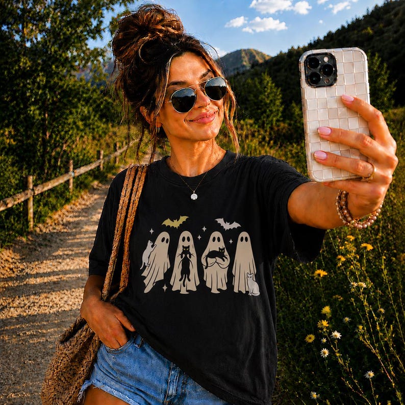 Pu&ograve; includere: Una t-shirt nera con una stampa grafica di quattro fantasmi beige, ognuno che interagisce con un gatto nero, accentuata da due piccoli pipistrelli e stelle. Indossata con pantaloncini di jeans blu e una borsa di paglia intrecciata.