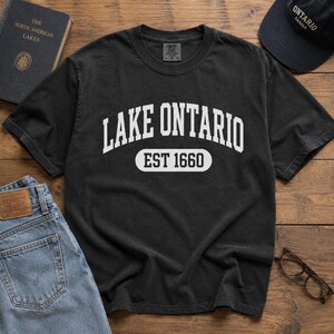 May include: A black short-sleeve t-shirt featuring the white text LAKE ONTARIO arched over the text EST 1660 inside a rounded rectangular border. The shirt has a crew neckline and a visible brand tag at the inner collar area.