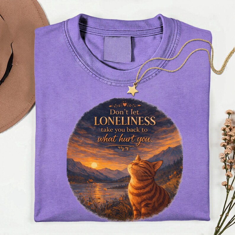 May include: A lavender t-shirt with a round graphic featuring a cat, lake, and sunset scene. The text reads "Don't let loneliness take you back to what hurt you." A gold star pendant necklace is draped on the shirt.