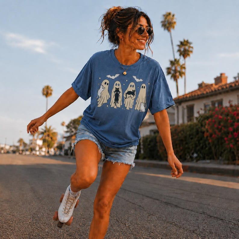 Pu&ograve; includere: Una t-shirt blu a maniche corte con una stampa grafica di quattro fantasmi che tengono in braccio dei gatti, accompagnata da piccole sagome di pipistrelli. La maglia ha un girocollo, una vestibilit&agrave; ampia, pantaloncini di jeans azzurri e un pattino bianco.