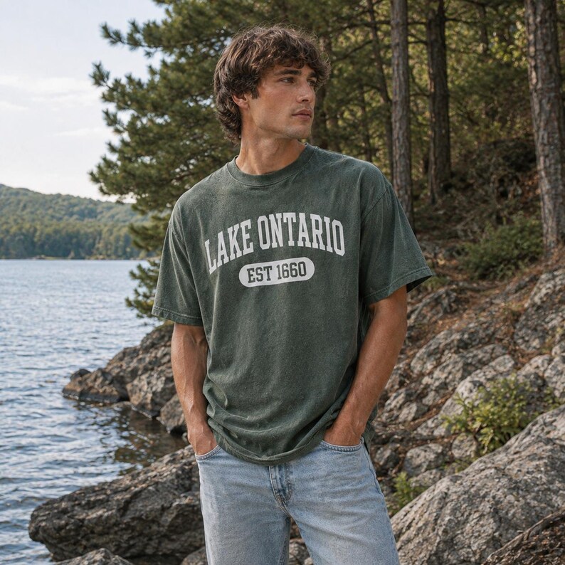 May include: A dark green short-sleeve t-shirt featuring the white text LAKE ONTARIO arched above a white oval containing the text EST 1660. The shirt has a crew neckline and a relaxed fit, paired with light blue denim jeans.