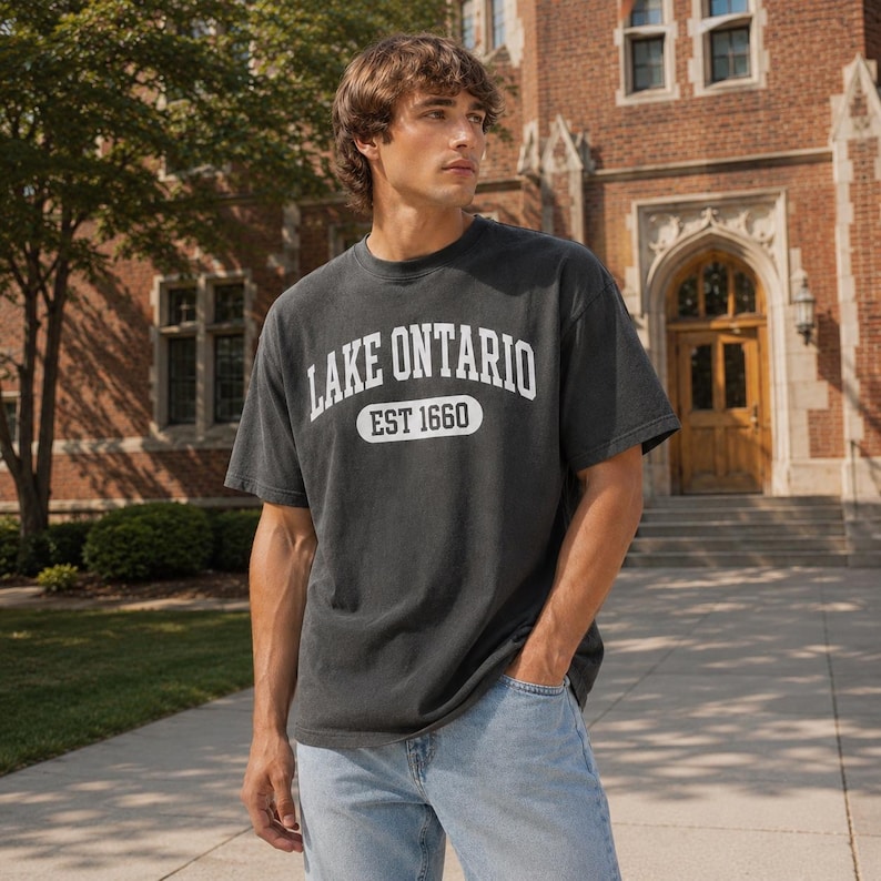 May include: A charcoal gray short-sleeve t-shirt featuring white text that reads LAKE ONTARIO and EST 1660 inside a white oval border. The shirt is paired with light blue denim jeans, creating a casual look with a classic collegiate style graphic.