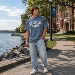 May include: A blue short-sleeved t-shirt featuring the white text LAKE ONTARIO and EST 1660 inside an oval graphic on the chest, paired with light blue denim jeans and white sneakers with grey accents, creating a casual outfit.