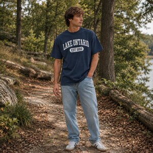 May include: A navy blue short-sleeved t-shirt with white text that reads LAKE ONTARIO EST 1660 across the chest, paired with light blue denim jeans and white sneakers with black stripes, worn in an outdoor setting with trees and a lake.