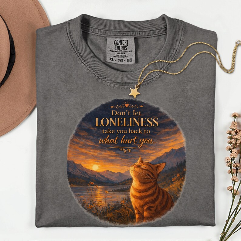 May include: A gray Comfort Colors t-shirt with a graphic of a cat and a sunset scene. The text on the shirt reads "Don't let LONELINESS take you back to what hurt you." A gold necklace with a star pendant is draped across the shirt.