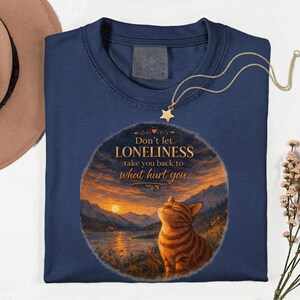 May include: Navy blue sweatshirt featuring a graphic of a cat gazing at a sunset over a lake and mountains. The text reads "Don't let loneliness take you back to what hurt you." A gold-colored necklace with a star pendant is draped over the shirt.