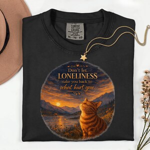 May include: Black Comfort Colors t-shirt with a graphic of a cat and a sunset scene. The text reads "Don't let LONELINESS take you back to what hurt you." A gold necklace with a star pendant is draped over the shirt.