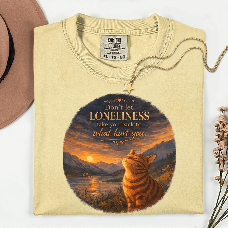 May include: A light yellow t-shirt with a graphic of a cat and a sunset scene. The text on the shirt reads "Don't let LONELINESS take you back to what hurt you." A gold-colored necklace with a star pendant is draped over the shirt.