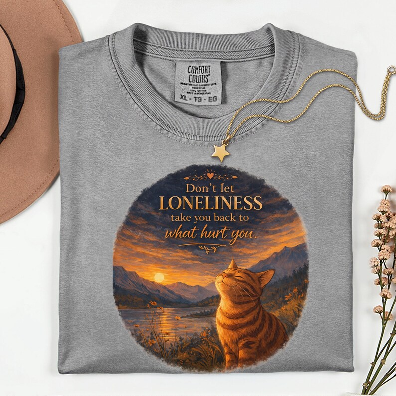 May include: A gray Comfort Colors XL-TG-EG t-shirt with a round graphic featuring a cat and the text "Don't let LONELINESS take you back to what hurt you." A gold star pendant necklace is draped over the shirt.