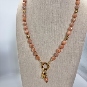 Puede incluir: Un collar de cuentas con piedras de sol facetadas en color melocotón intercaladas con pequeños espaciadores de metal dorado. El collar incluye un cierre circular dorado central con un pequeño dije de cruz dorada y una cuenta a juego.