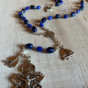 May include: A rosary with blue beads and silver accents. The beads vary in shade from dark navy to lighter blue. The rosary features a silver crucifix and sailboat charms. The background is a light-colored wooden surface.