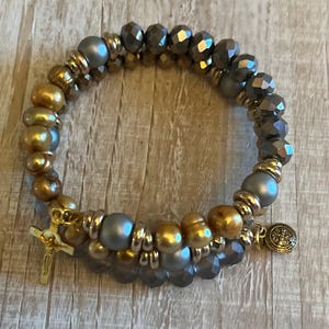May include: A beaded bracelet featuring a mix of gold, gray, and faceted beads. The bracelet includes a gold cross charm and a round charm with a religious emblem. The beads are strung together with elastic.