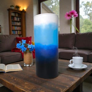May include: A tall, cylindrical tumbler with a gradient design, transitioning from white at the top to shades of blue and ending in black at the base. The tumbler is on a wooden table, with a cup of coffee and flowers in the background.
