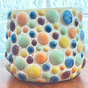 Könnte beinhalten: Ein Keramik-Pflanzgefäß mit einer strukturierten Oberfläche, die mit bunten, erhabenen, blasenartigen Formen bedeckt ist. Die Blasen sind in den Farbtönen Blau, Grün, Orange, Gelb und Lila gehalten. Der Pflanztopf steht auf einer Holzoberfläche.