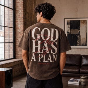 A person wearing a brown t-shirt with the text "GOD HAS A PLAN" printed on it, standing in a room with a brick wall and a window.