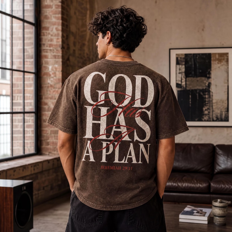 A person wearing a brown t-shirt with the text "GOD HAS A PLAN" printed on it, standing in a room with a brick wall and a window.