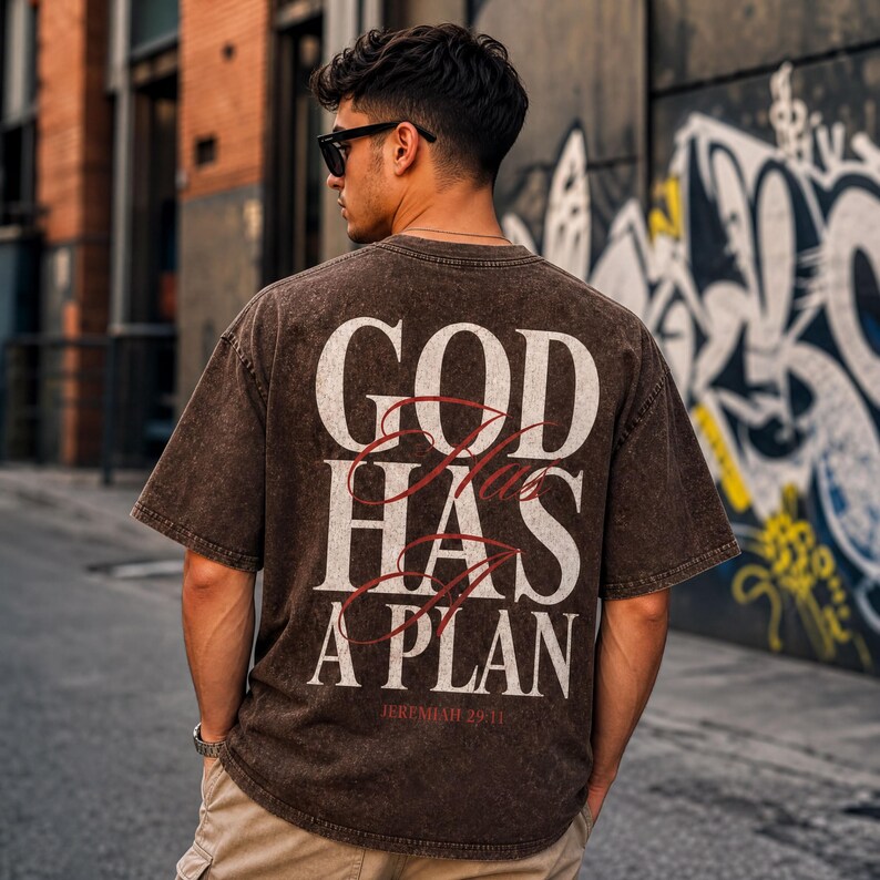 A man wearing a brown t-shirt with the text "GOD HAS A PLAN" printed on it, standing in front of a graffiti-covered wall.