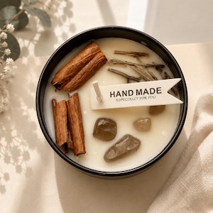 May include: A round black candle container filled with white wax, topped with two cinnamon sticks, three small brown stones, thin wooden twigs, and a white flag-shaped tag with the text HAND MADE ESPECIALLY FOR YOU printed in black.