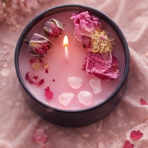 May include: A lit pink candle in a black container, adorned with dried rose buds, petals, and crystals. The candle's surface is decorated with dried flowers and rose petals, creating a romantic and natural aesthetic. The candle is set on a pink fabric.