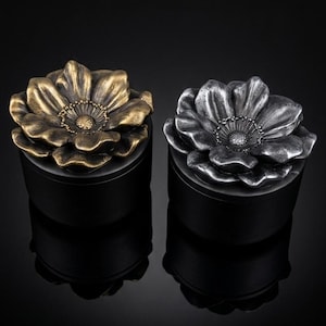 May include: Two decorative boxes with floral lids. One lid is gold-toned, the other is silver-toned. Both sit atop black cylindrical bases. The background is a dark, reflective surface.