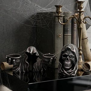 Może przedstawiać: Wystawa o tematyce Halloween z dwoma ciemnymi metalowymi świecznikami w kształcie czaszki i świecznikiem w kształcie postaci w kapturze. Ekspozycja obejmuje antyczne książki, mosiężny świecznik i sztuczne pajęczyny.