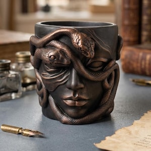 Könnte beinhalten: Ein zylindrischer, dunkelgrauer Behälter mit einem skulpturalen menschlichen Gesicht im Relief, verziert mit mehreren ineinander verschlungenen Schlangen in metallischem Bronze-Finish, die sich um die Augen und Wangen winden und eine texturierte Oberfläche bilden.