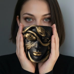Pode incluir: Uma caneca de cerâmica preta com um design de Medusa, apresentando cobras e traços faciais dourados. A caneca é segurada em frente ao rosto de uma pessoa, mostrando os detalhes intrincados da obra de arte. O design da caneca é uma mistura marcante de tons escuros e dourados.