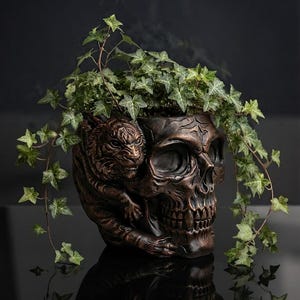 May include: A dark bronze-coloured planter shaped like a human skull with a tiger sculpted onto the side. The skull features deep eye sockets and a detailed jaw. A green ivy plant grows out of the top of the skull, with vines draping over the sides.