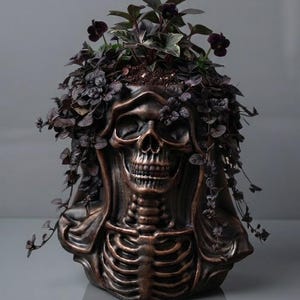 May include: A bronze-colored planter shaped like a human skull and upper torso, featuring a hooded design. The skull is filled with a dark-leafed plant that spills over the sides, creating a dense, cascading effect around the skeletal structure.