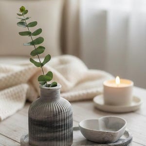 May include: A serene still life featuring a small, ribbed vase with a sprig of greenery, a heart-shaped bowl, and a lit candle. The items rest on a marble tray, creating a calming aesthetic. The scene is set on a light-colored wooden surface.