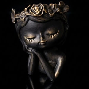 May include: A dark bronze-colored decorative planter in the shape of a woman's head and shoulders. The woman is depicted with closed eyes, a serene expression, and a floral crown. The planter is resting on a reflective surface.
