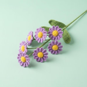Op de afbeelding: Een gehaakte bloemstengel met meerdere paarse madeliefachtige bloemen met gele centra en groene bladeren. De handgemaakte bloemen zijn gerangschikt op een lichtgroene achtergrond. Het handwerk is gedetailleerd en delicaat.