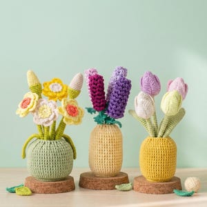 Puede incluir: Tres arreglos florales de ganchillo en jarrones. El primer jarrón es verde y contiene flores amarillas y blancas con centros naranjas. El segundo jarrón es amarillo y contiene flores moradas y rosas. El tercer jarrón es amarillo y contiene tulipanes morados y blancos.