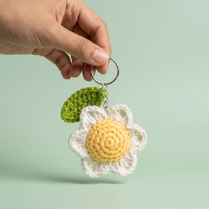 May include: A crochet flower keychain featuring a central yellow circular disc surrounded by six white petals and a single green leaf attached to the top. The flower is connected to a silver metal split ring by a short silver metal chain.