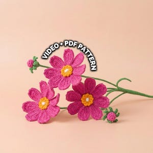 May include: Crocheted pink flowers with yellow centers and green stems. The flowers are arranged on a light peach background. The text "VIDEO + PDF PATTERN" is arched above the flowers.
