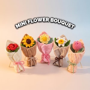 Puede incluir: Cinco pequeños ramos de flores de ganchillo. Cada ramo presenta una flor diferente, incluyendo una rosa roja, un girasol, una margarita, un narciso y un tulipán. Los ramos están envueltos en varios colores de hilo y atados con cintas.