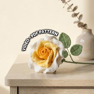 May include: A handcrafted crochet rose with white and yellow petals, green leaves, and a stem. The text "VIDEO * PDF PATTERN" is displayed on the left side of the rose. The rose is placed on a wooden surface.