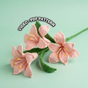 Puede incluir: Primer plano de tres flores de lirio rosas tejidas con ganchillo con tallos y hojas verdes. Las flores tienen estambres amarillos. El texto "VIDEO + PDF PATTERN" está en la esquina superior izquierda.