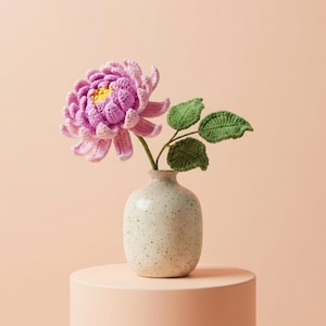 Könnte beinhalten: Eine handgefertigte Häkelblume in einer gesprenkelten Vase. Die Blume ist ein leuchtendes Pink mit gelber Mitte und grünen Blättern. Die Vase ist cremefarben und steht auf einem hellrosa Sockel, was eine weiche, einladende Ästhetik erzeugt.