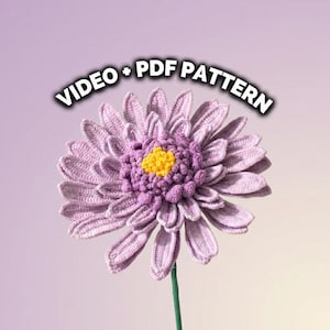 Op de afbeelding: Een handgemaakte gehaakte bloem in tinten lavendel en paars, met een heldergeel centrum. De bloem heeft gedetailleerd bloemblaadjeswerk en een groene stengel. De tekst bovenaan luidt "VIDEO + PDF PATTERN".