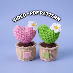 May include: Two crocheted heart-shaped cacti in tan pots with brown soil tops. One cactus is pink, the other is green, and both feature a white flower with a yellow centre. Each pot has a small tag labelled HAND MADE. Text reads VIDEO + PDF PATTERN.