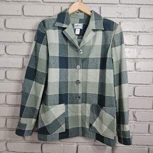 Könnte beinhalten: Ein grün-blau karierter Blazer mit Kragen, drei Knöpfen und zwei Vordertaschen. Der Blazer hängt an einem Holzbügel vor einer Backsteinmauer.