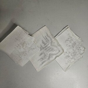 May include: Three white handkerchiefs with embroidered designs. One features butterflies, another a geometric pattern, and the third floral motifs. The handkerchiefs are folded and arranged on a light gray surface.