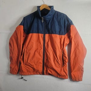 May include: A color-blocked jacket featuring a dark blue upper section and a vibrant orange lower section. This long-sleeved garment includes a full-length front zipper, a high collar, and two side pockets with zippers, finished with an adjustable hem.