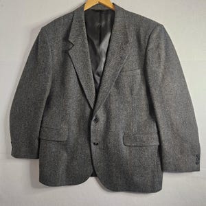 May include: A charcoal gray blazer featuring a subtle herringbone pattern, a notched lapel, and a two-button front closure. The garment includes two flap pockets at the waist, a single chest pocket, and a smooth black interior lining.