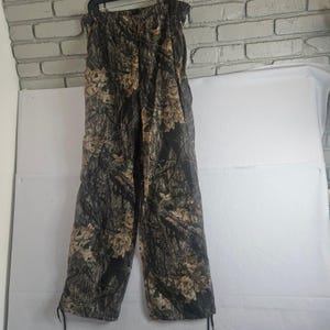 May include: Full-length trousers featuring an all-over camouflage pattern in shades of dark brown, black, and tan. The pants have an elasticized waistband and adjustable drawstrings at the bottom of each leg hem for a customizable fit.