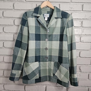 May include: A green and blue plaid blazer with a collared neckline, three buttons, and two front pockets. The blazer is hanging on a wooden hanger against a brick wall.