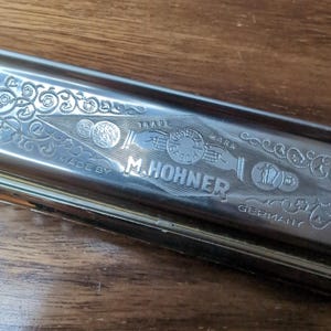 以下が含まれることがあります： 複雑な花の彫刻とM. HOHNER、TRADE MARK、GESETZ、GESCH、MADE BY、GERMANYの文字が刻まれた銀色の金属製ハーモニカ。金属製のケースの表面には、手、コイン、ハープの詳細なイラストが描かれています。