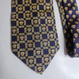 Pode incluir: Uma gravata azul-marinho escuro com um padrão repetitivo dourado do logótipo da cabeça da Medusa, inserido numa borda quadrada com padrão grego. O design é consistente em todo o tecido, exibindo uma grelha geométrica de motivos dourados.